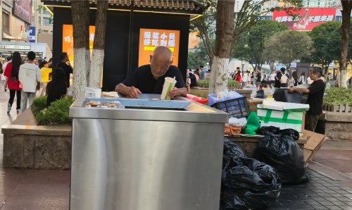 Poor Chinese man looking through a bin in a busy town square.